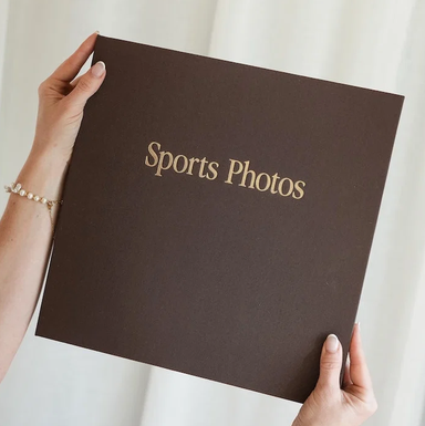 A person holds a large dark brown sports photo album with gold foil lettering that reads 'Sports Photos' against a soft white curtain background.