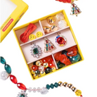 A colorful beaded jewelry set displayed in an open yellow compartment box, with matching loose beaded strands draped around it on a white background. The set includes red, green, gold, and clear beads with decorative charms, and a yellow box is partially visible at the top left.