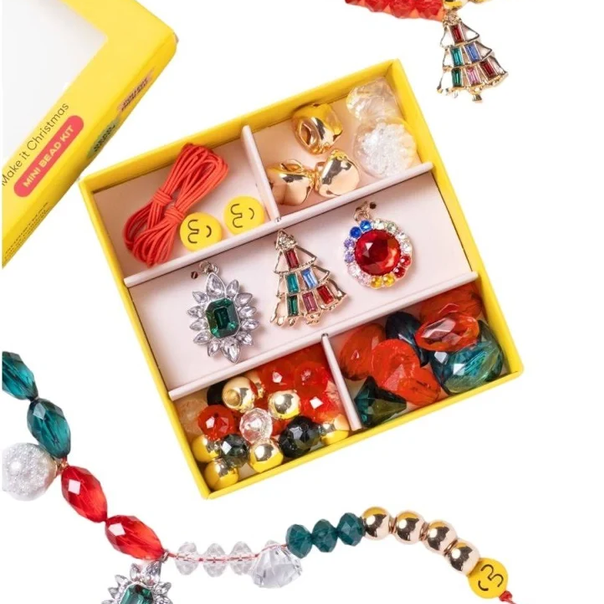 A colorful beaded jewelry set displayed in an open yellow compartment box, with matching loose beaded strands draped around it on a white background. The set includes red, green, gold, and clear beads with decorative charms, and a yellow box is partially visible at the top left.