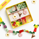 A colorful DIY craft kit in a yellow box with compartments containing beads, charms, and gummy-like pieces, surrounded by completed fruit and gummy bead garlands on a white background.